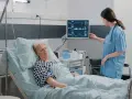 Enfermera revisa monitor de paciente en cama. El monitor muestra constantes vitales, tcae que es un signo de recuperación.