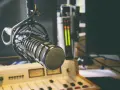 Micrófono profesional en estudio, listo para la comunicación audiovisual. Salidas de audio y consola de mezcla en el fondo.