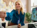 Mujer sonríe mientras da la mano, con un mapa mundial en la pared y en el monitor. Un buen sueldo en negocios internacionales es posible.