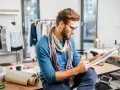 Un diseñador de producto con gafas y bufanda revisa bocetos de moda en su taller, rodeado de maniquíes y telas.