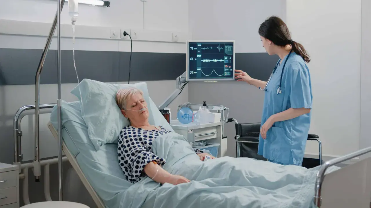 Enfermera revisa monitor de paciente en cama. El monitor muestra constantes vitales, tcae que es un signo de recuperación.