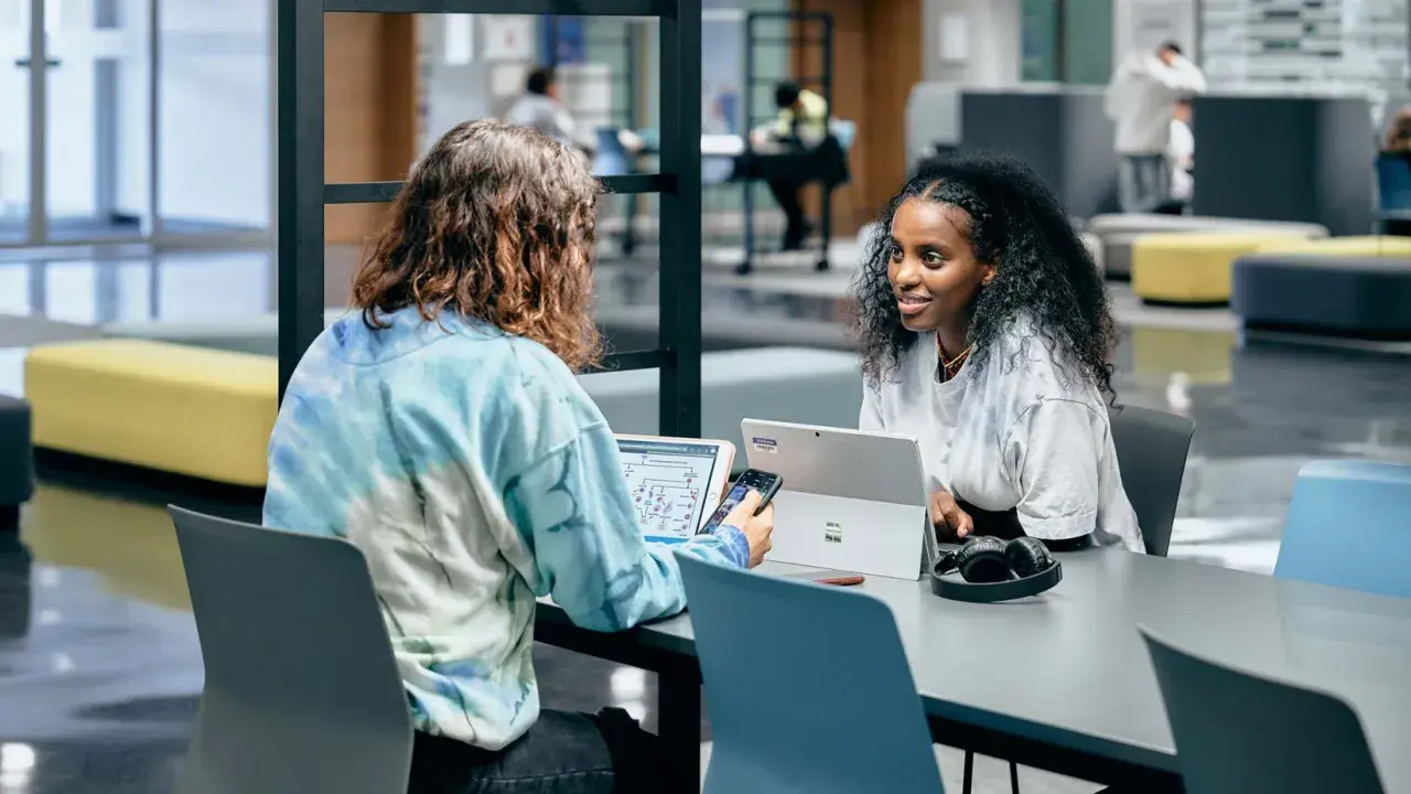 Dos estudiantes colaboran en un proyecto, discutiendo ideas mientras trabajan en sus laptops. Su trabajo en equipo es un ejemplo de cómo el **trabajo social** puede fomentar el aprendizaje y la innovación.