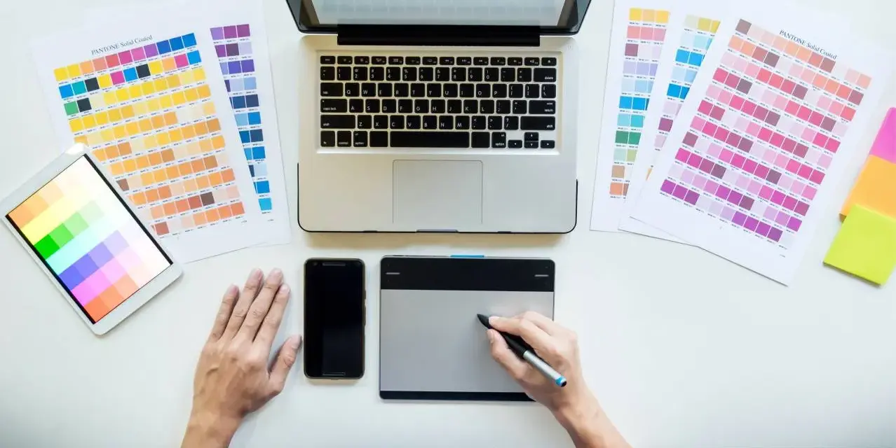 Diseñador gráfico trabajando en su escritorio con paletas de colores, tablet, móvil y portátil. Así se ve el día a día de cuanto gana un diseñador gráfico.