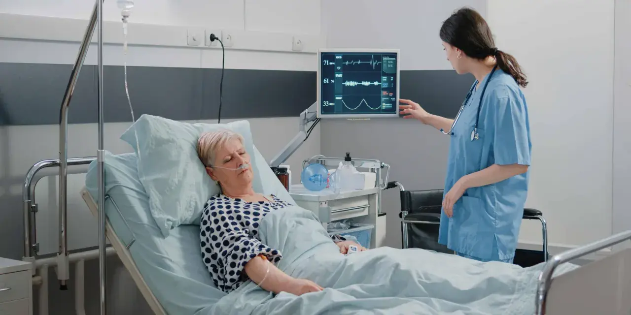 Enfermera revisa monitor de paciente en cama. El monitor muestra constantes vitales, tcae que es un signo de recuperación.