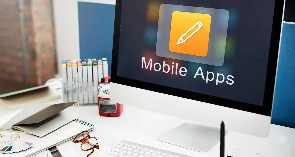 Escritorio de diseñador con ordenador mostrando "Mobile Apps", lápices de colores, gafas y cuadernos.