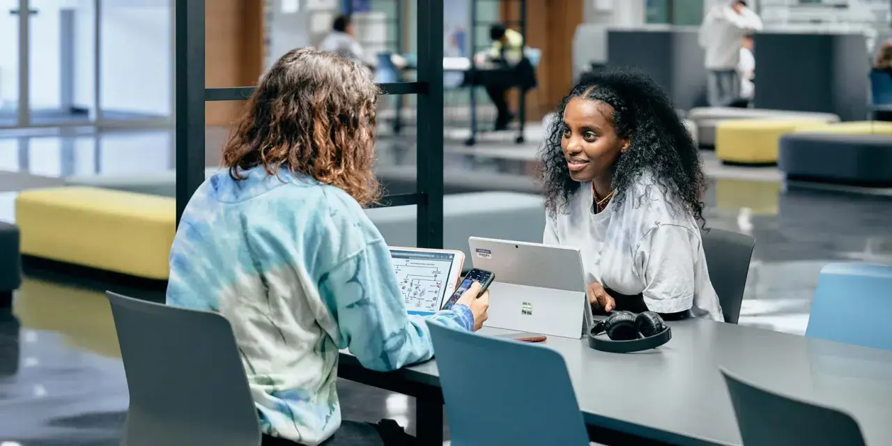 Dos estudiantes colaboran en un proyecto, discutiendo ideas mientras trabajan en sus laptops. Su trabajo en equipo es un ejemplo de cómo el **trabajo social** puede fomentar el aprendizaje y la innovación.