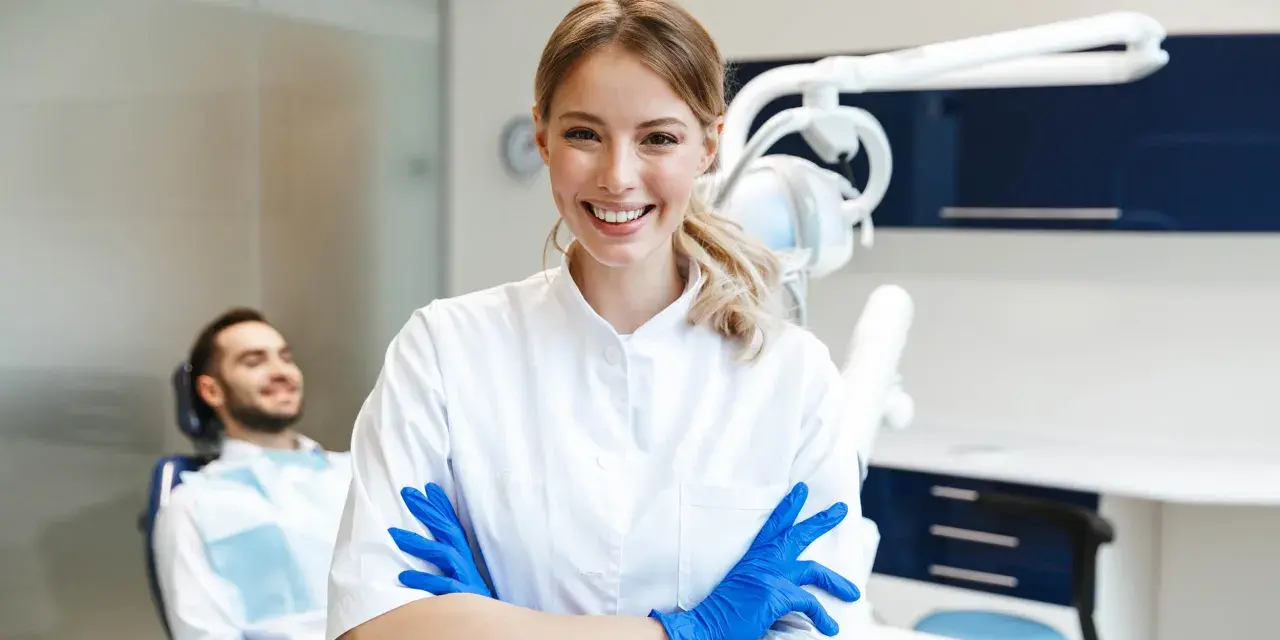 Sonrisa de profesional dental con guantes azules, lista para impartir un curso de higiene bucodental. Paciente relajado en silla dental al fondo.