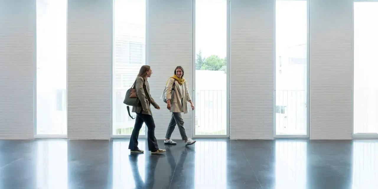 Dos estudiantes caminan por un pasillo moderno, quizás en una de las **biomedicina universidades**, con grandes ventanales y paredes de ladrillo blanco.