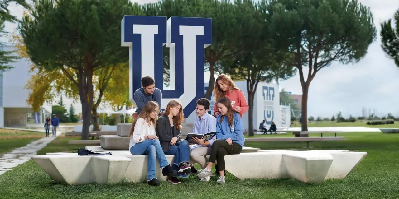Estudiantes discuten en el campus de la Universidad Francisco de Vitoria, rodeados de árboles y el logo de la institución. Buenas **universidad francisco de vitoria opiniones** para elegir tu futuro.