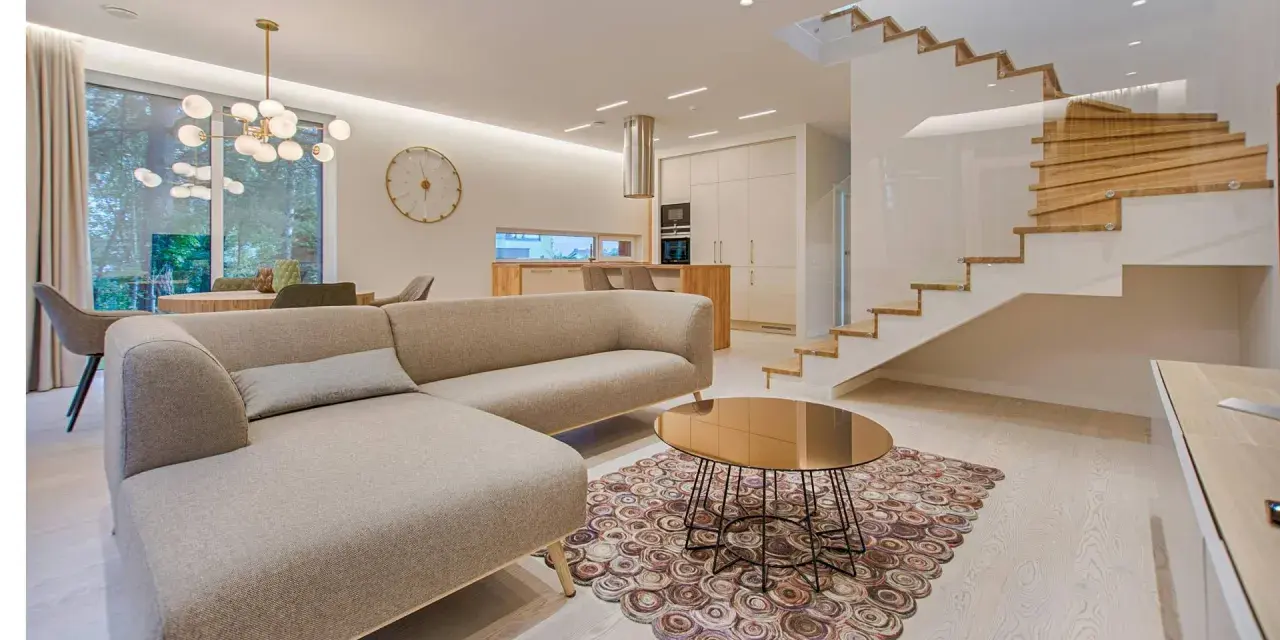 Elegante salón con sofá beige, mesa de centro dorada y alfombra circular. Escalera moderna de madera y cristal. La diferencia entre decorador y diseñador de interiores se refleja en la armonía del espacio.