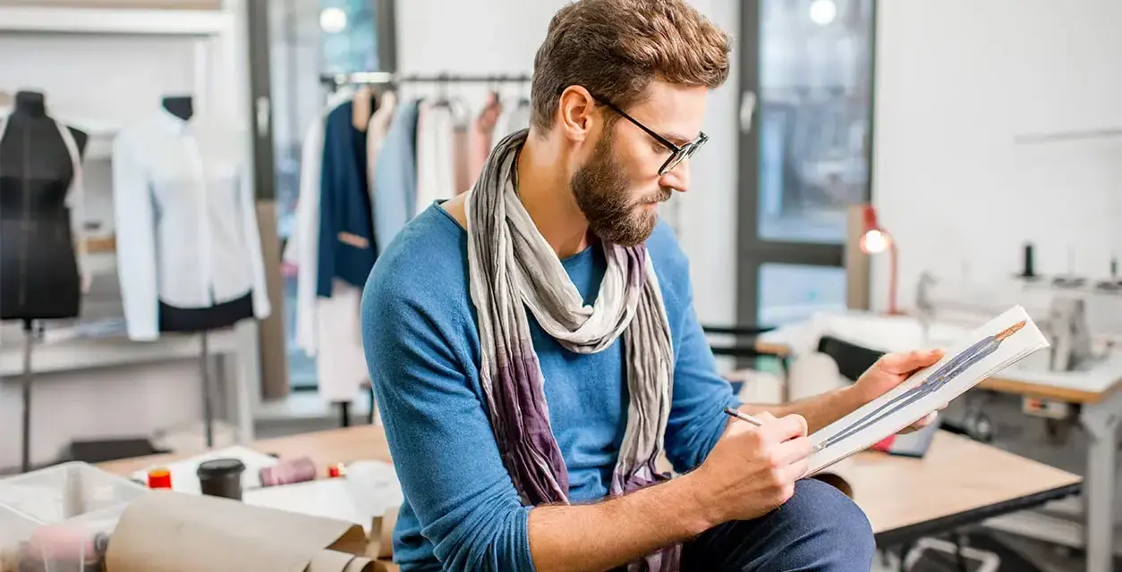 Un diseñador de producto con gafas y bufanda revisa bocetos de moda en su taller, rodeado de maniquíes y telas.