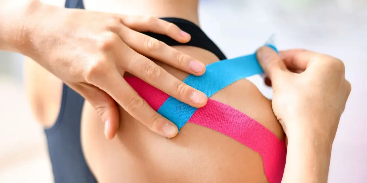 Manos aplican cinta kinesiológica en un hombro. Una carrera en fisioterapia te enseña estas técnicas.