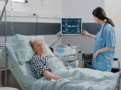 Enfermera revisa monitor de paciente en cama. El monitor muestra constantes vitales, tcae que es un signo de recuperación.