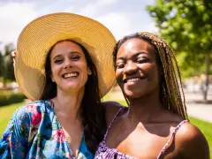 Dos amigas sonriendo bajo el sol, una con sombrero y vestido floral, la otra con trenzas. Una imagen personal de alegría compartida.