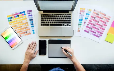 Diseñador gráfico trabajando en su escritorio con paletas de colores, tablet, móvil y portátil. Así se ve el día a día de cuanto gana un diseñador gráfico.