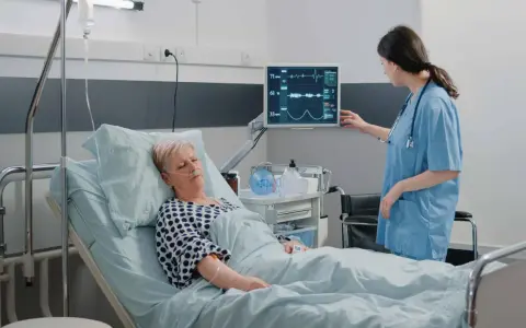 Enfermera revisa monitor de paciente en cama. El monitor muestra constantes vitales, tcae que es un signo de recuperación.