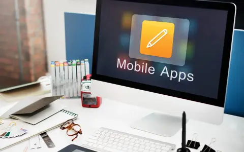 Escritorio de diseñador con ordenador mostrando "Mobile Apps", lápices de colores, gafas y cuadernos.
