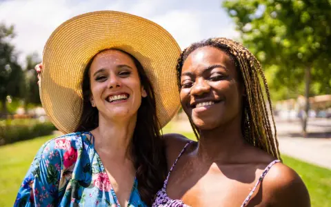 Dos amigas sonriendo bajo el sol, una con sombrero y vestido floral, la otra con trenzas. Una imagen personal de alegría compartida.