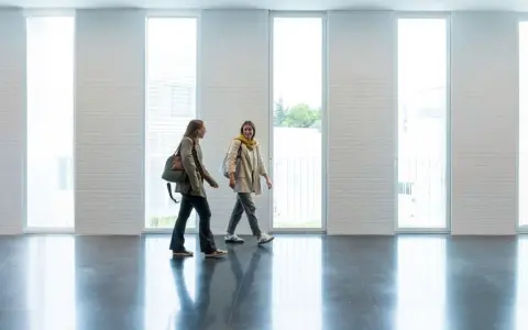 Dos estudiantes caminan por un pasillo moderno, quizás en una de las **biomedicina universidades**, con grandes ventanales y paredes de ladrillo blanco.