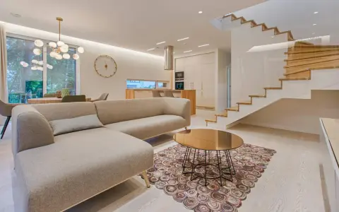 Elegante salón con sofá beige, mesa de centro dorada y alfombra circular. Escalera moderna de madera y cristal. La diferencia entre decorador y diseñador de interiores se refleja en la armonía del espacio.