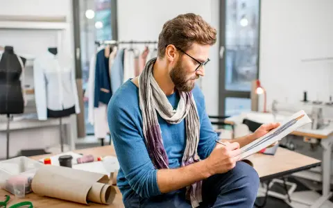 Un diseñador de producto con gafas y bufanda revisa bocetos de moda en su taller, rodeado de maniquíes y telas.