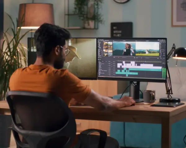 Hombre trabajando en la edición de video profesional en su estudio casero, con dos monitores mostrando la línea de tiempo y clips.