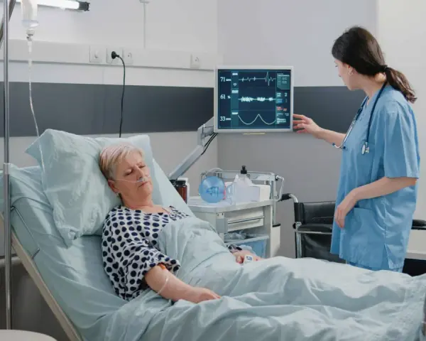 Enfermera revisa monitor de paciente en cama. El monitor muestra constantes vitales, tcae que es un signo de recuperación.