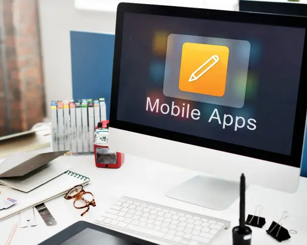 Escritorio de diseñador con ordenador mostrando "Mobile Apps", lápices de colores, gafas y cuadernos.
