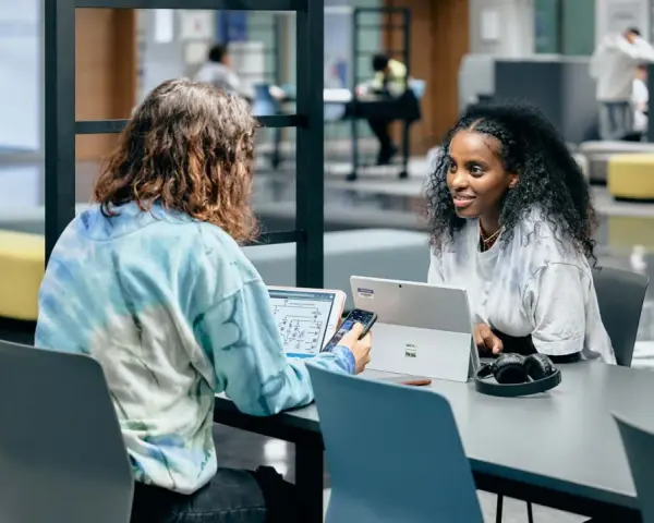 Dos estudiantes colaboran en un proyecto, discutiendo ideas mientras trabajan en sus laptops. Su trabajo en equipo es un ejemplo de cómo el **trabajo social** puede fomentar el aprendizaje y la innovación.