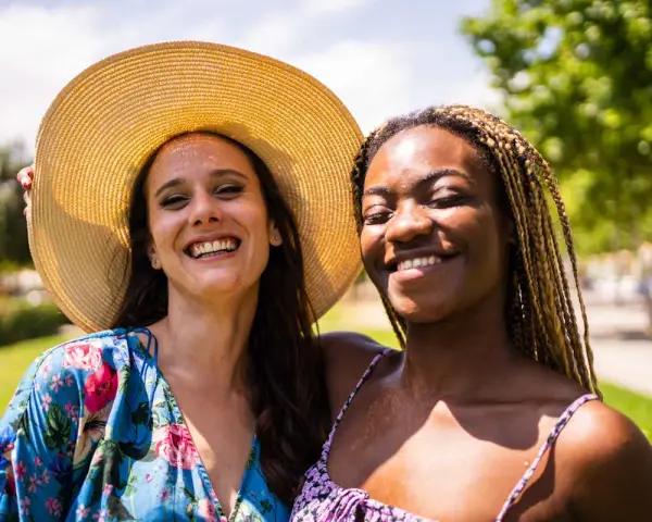 Dos amigas sonriendo bajo el sol, una con sombrero y vestido floral, la otra con trenzas. Una imagen personal de alegría compartida.