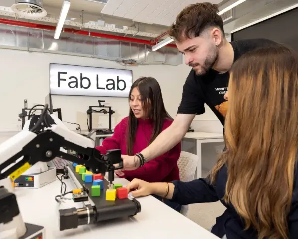Estudiantes en un Fab Lab trabajan con un brazo robótico y bloques de colores, explorando la automatización y las relaciones internacionales.