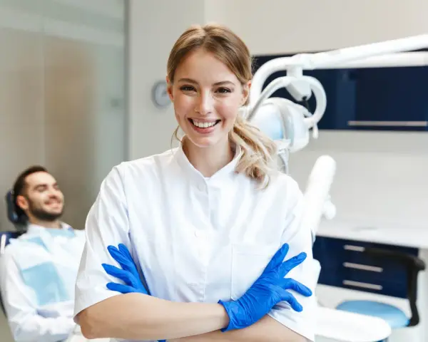 Sonrisa de profesional dental con guantes azules, lista para impartir un curso de higiene bucodental. Paciente relajado en silla dental al fondo.