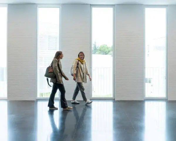 Dos estudiantes caminan por un pasillo moderno, quizás en una de las **biomedicina universidades**, con grandes ventanales y paredes de ladrillo blanco.