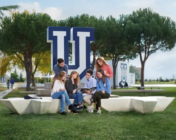 Estudiantes discuten en el campus de la Universidad Francisco de Vitoria, rodeados de árboles y el logo de la institución. Buenas **universidad francisco de vitoria opiniones** para elegir tu futuro.