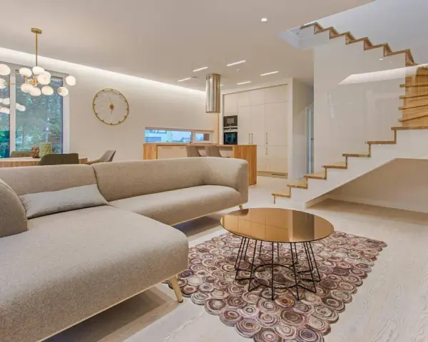 Elegante salón con sofá beige, mesa de centro dorada y alfombra circular. Escalera moderna de madera y cristal. La diferencia entre decorador y diseñador de interiores se refleja en la armonía del espacio.