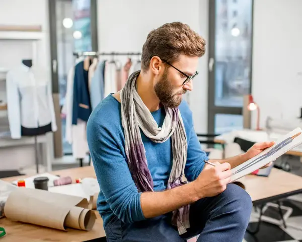 Un diseñador de producto con gafas y bufanda revisa bocetos de moda en su taller, rodeado de maniquíes y telas.