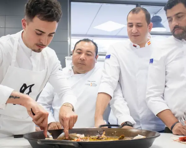 Cuatro chefs observan atentamente una paella. Se debate la diferencia entre chef y cocinero mientras preparan el plato.