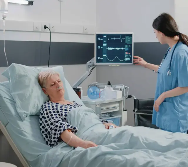 Enfermera revisa monitor de paciente en cama. El monitor muestra constantes vitales, tcae que es un signo de recuperación.