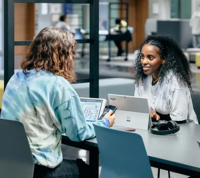 Dos estudiantes colaboran en un proyecto, discutiendo ideas mientras trabajan en sus laptops. Su trabajo en equipo es un ejemplo de cómo el **trabajo social** puede fomentar el aprendizaje y la innovación.