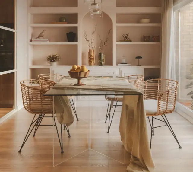 Comedor moderno con mesa de cristal y sillas de ratán. Estanterías empotradas y un toque de diseño de interiores cálido.