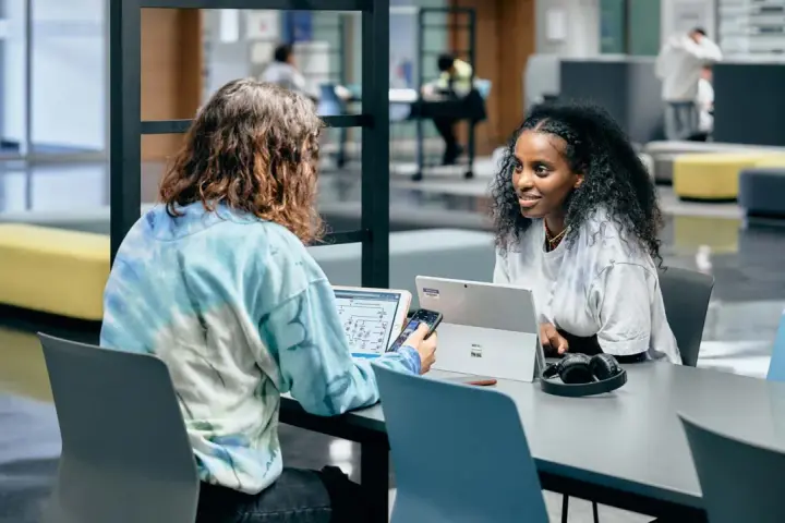 Dos estudiantes colaboran en un proyecto, discutiendo ideas mientras trabajan en sus laptops. Su trabajo en equipo es un ejemplo de cómo el **trabajo social** puede fomentar el aprendizaje y la innovación.