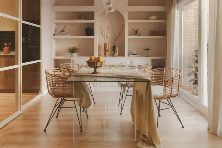 Comedor moderno con mesa de cristal y sillas de ratán. Estanterías empotradas y un toque de diseño de interiores cálido.