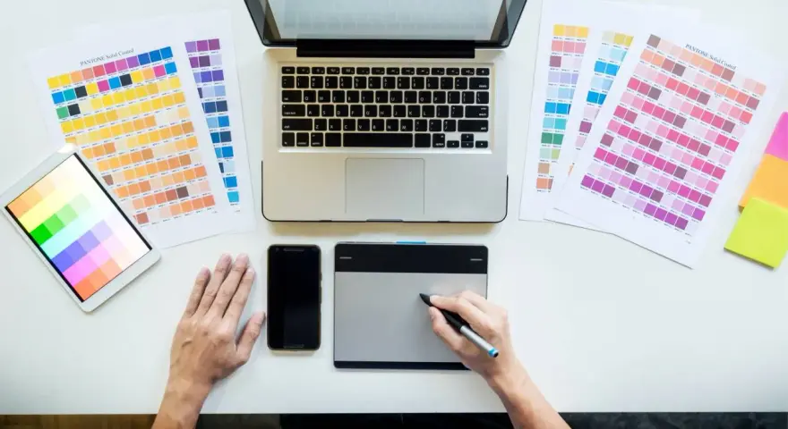 Diseñador gráfico trabajando en su escritorio con paletas de colores, tablet, móvil y portátil. Así se ve el día a día de cuanto gana un diseñador gráfico.
