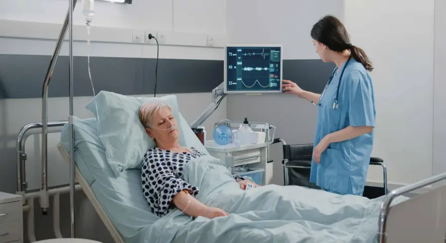 Enfermera revisa monitor de paciente en cama. El monitor muestra constantes vitales, tcae que es un signo de recuperación.