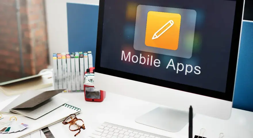 Escritorio de diseñador con ordenador mostrando "Mobile Apps", lápices de colores, gafas y cuadernos.
