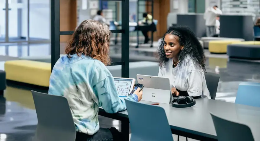Dos estudiantes colaboran en un proyecto, discutiendo ideas mientras trabajan en sus laptops. Su trabajo en equipo es un ejemplo de cómo el **trabajo social** puede fomentar el aprendizaje y la innovación.
