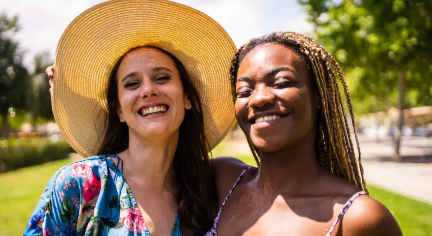 Dos amigas sonriendo bajo el sol, una con sombrero y vestido floral, la otra con trenzas. Una imagen personal de alegría compartida.