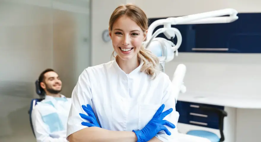 Sonrisa de profesional dental con guantes azules, lista para impartir un curso de higiene bucodental. Paciente relajado en silla dental al fondo.