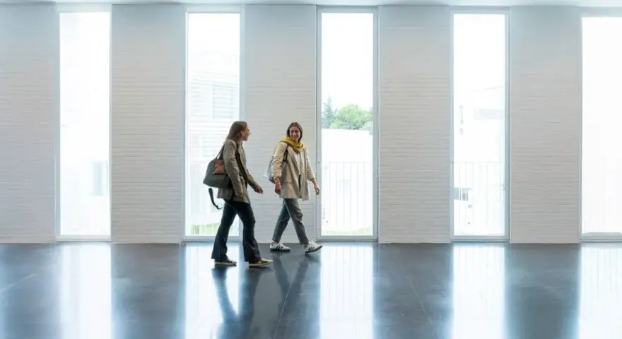 Dos estudiantes caminan por un pasillo moderno, quizás en una de las **biomedicina universidades**, con grandes ventanales y paredes de ladrillo blanco.