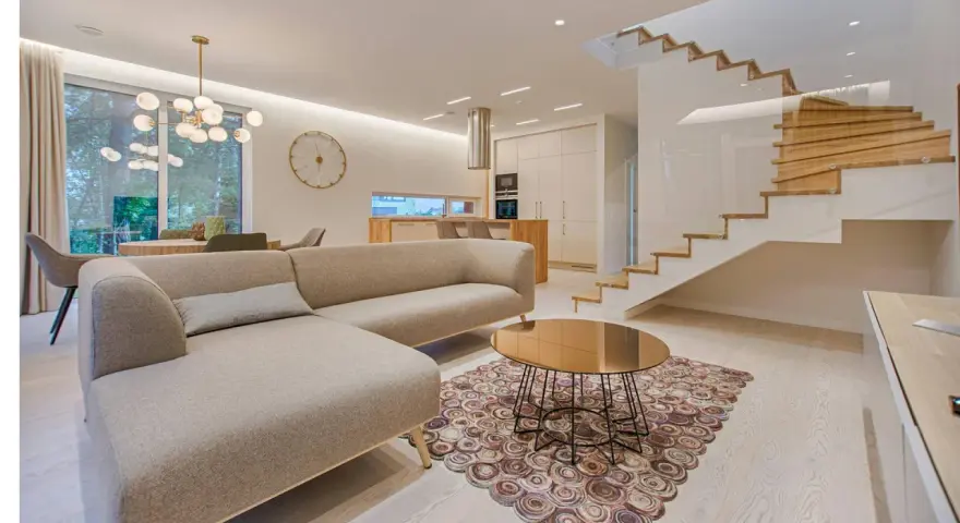 Elegante salón con sofá beige, mesa de centro dorada y alfombra circular. Escalera moderna de madera y cristal. La diferencia entre decorador y diseñador de interiores se refleja en la armonía del espacio.